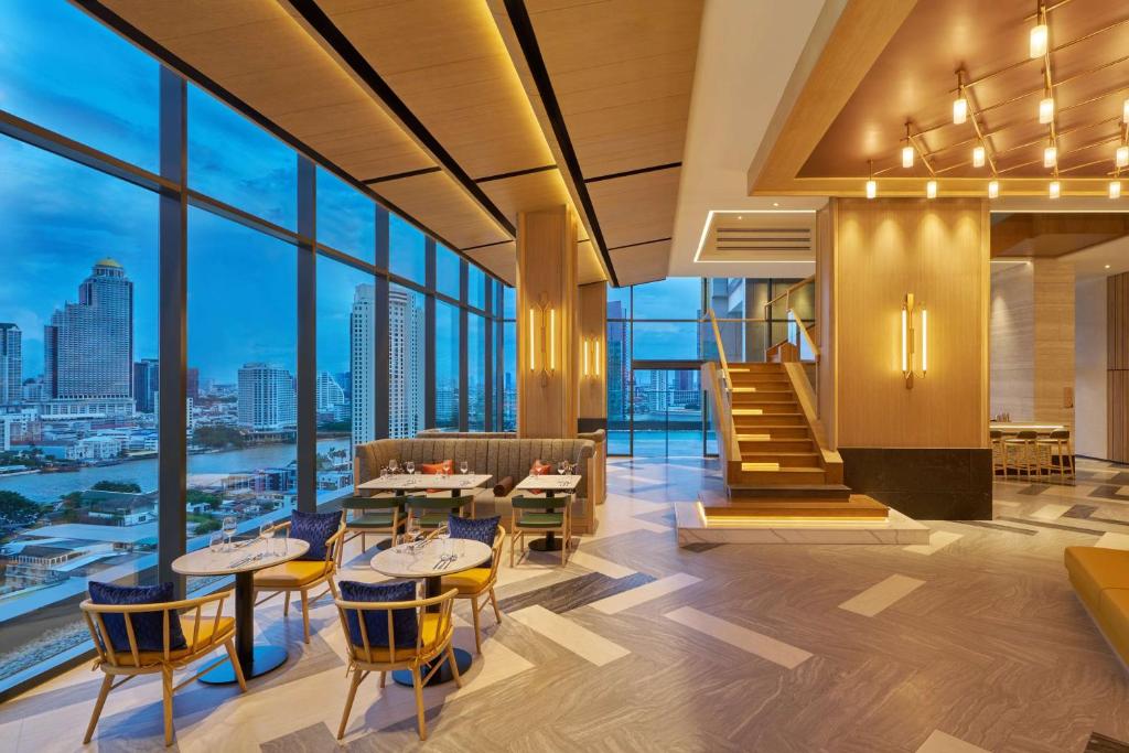 A bright lounge overlooks Bangkok through tall windows, with café tables, armchairs, and a staircase.