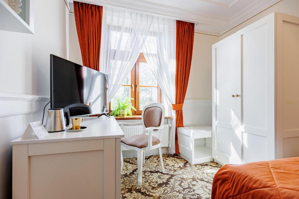 Sunlit desk by window with TV, kettle, white chair and orange curtains, patterned carpet.