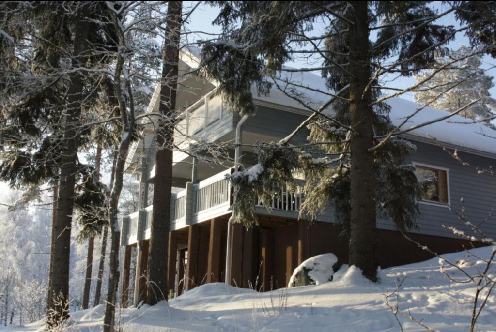 una casa cubierta de nieve con árboles en el primer plano en Tahkon Riihiranta - 4 bedroom villa with lake view - Tilava mökki rantanäkymin, en Tahkovuori