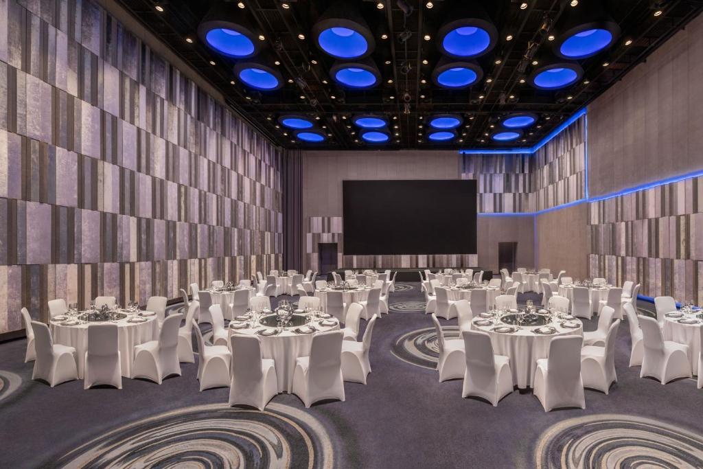 A large banquet hall set with round tables, white chairs and blue accent lighting around a presentation screen.