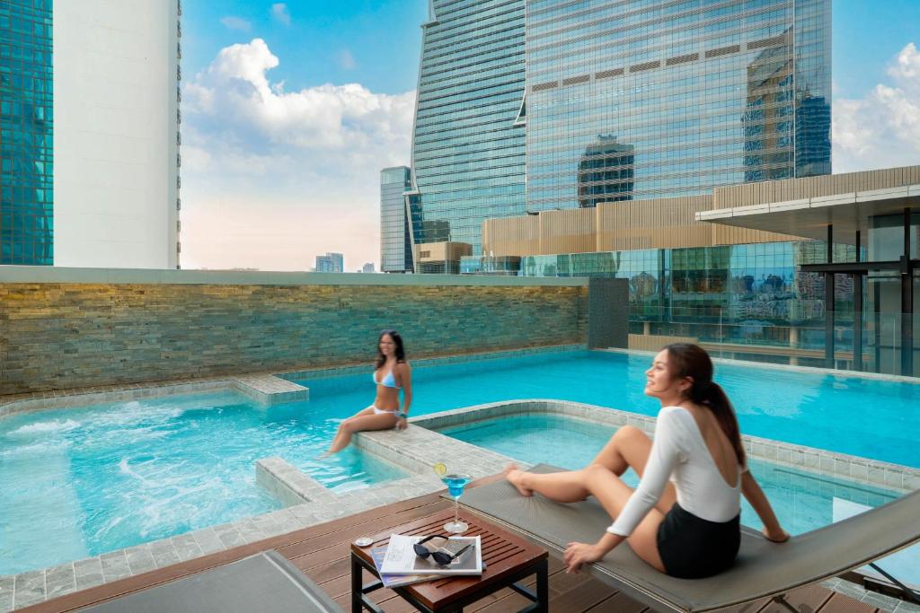 A rooftop pool deck with loungers, a shallow ledge and a high-rise city backdrop.