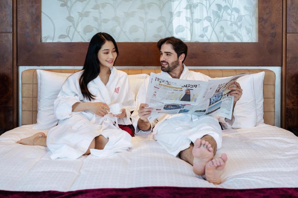 a man and woman sitting on a bed reading a newspaper at Grand Millennium Al Wahda Hotel and Executive Apartments Abu Dhabi in Abu Dhabi