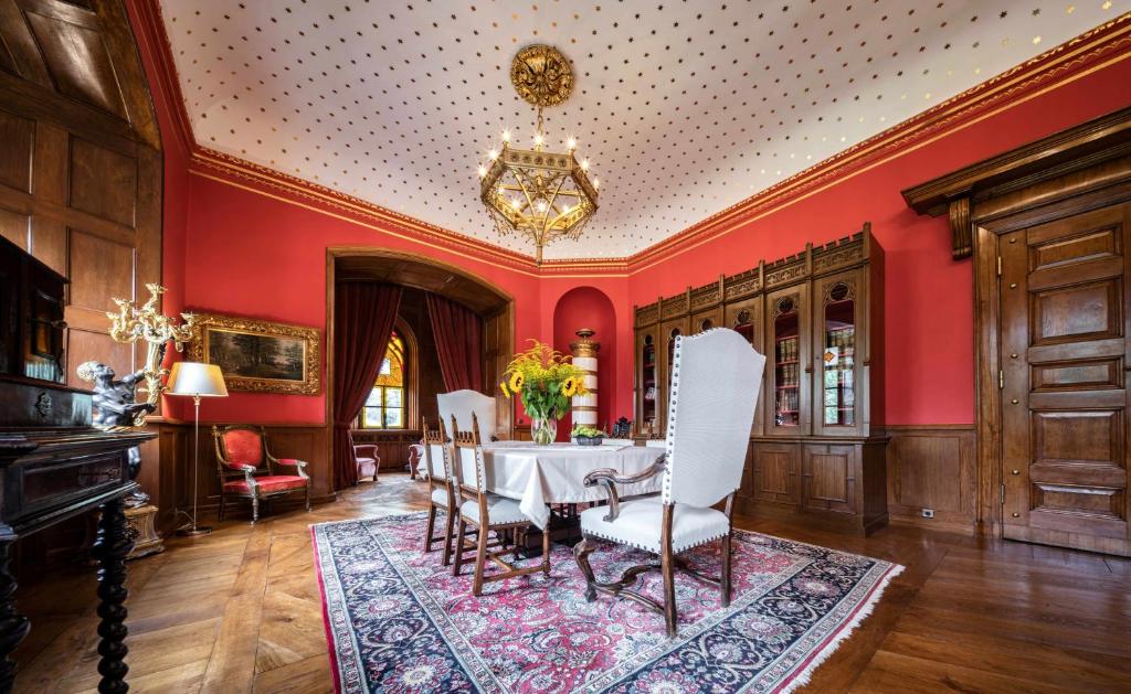 Formal dining room with red walls, carved wood paneling, long table, white chairs, ornate chandelier.