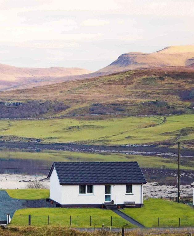 a white house in a field with mountains in the background at Almondbank Cottage 8 miles from Portree 2 bedrooms sleeps 3 adults in Skeabost