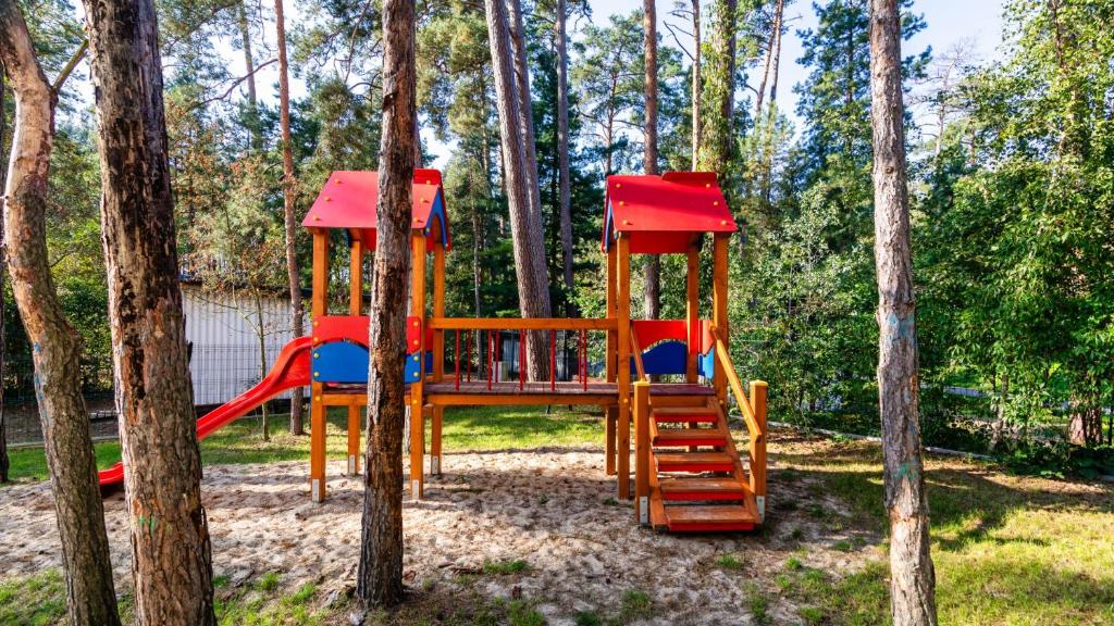 Wooden children's playground with red-roofed towers, slide and bridge, nestled among tall pine trees.