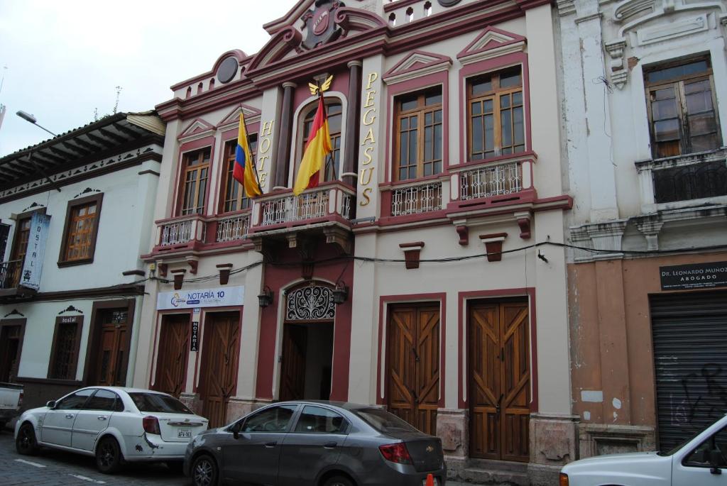 dos autos estacionados frente a un edificio con banderas. en Hotel Pegasus, en Cuenca