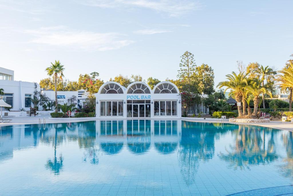 a swimming pool with a building and palm trees at Iris Hotel & Thalasso in Taguermess