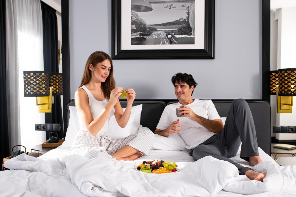 a man and woman sitting on a bed eating food at Athenian Riviera Hotel& Suites in Athens