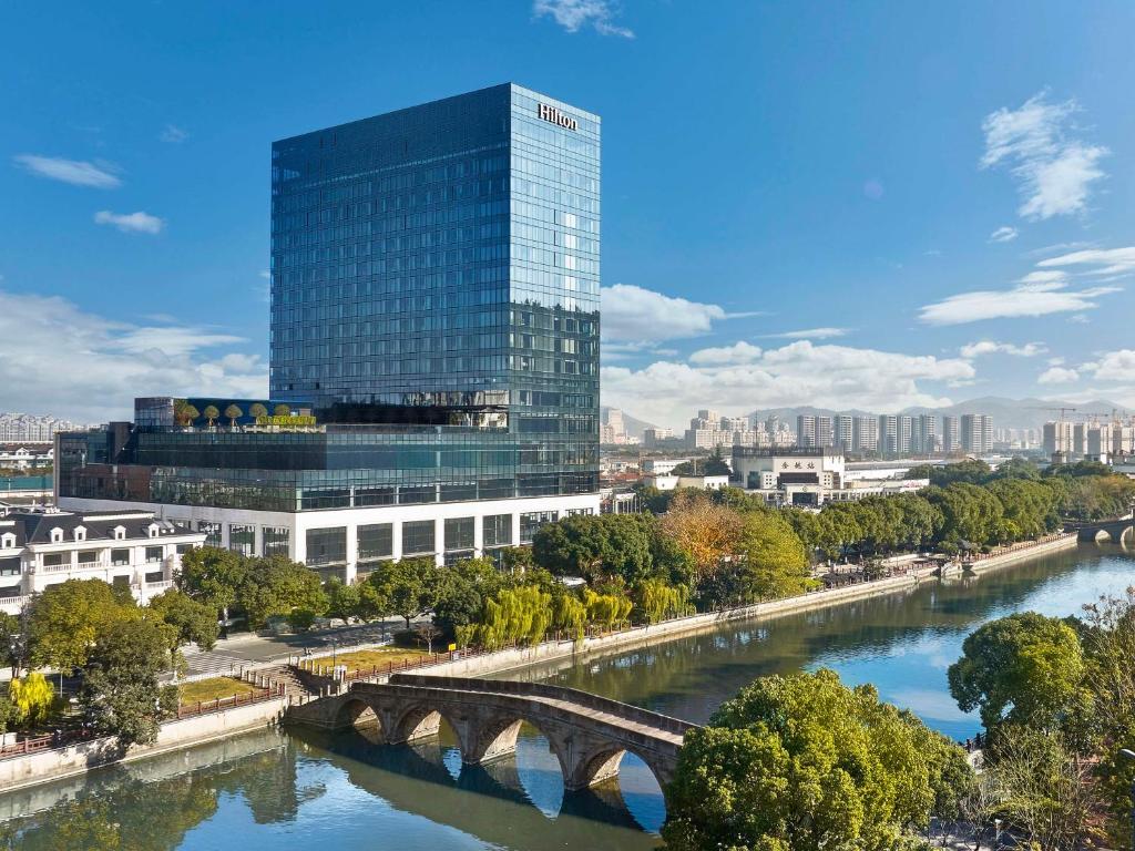 a tall glass building next to a river with a bridge at Hilton Yuyao in Yuyao