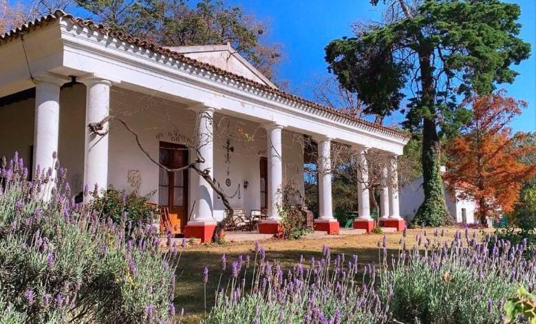 un bâtiment blanc avec des colonnes blanches et des fleurs violettes dans l'établissement Estancia Pozo de Correa, à Agua de las Piedras