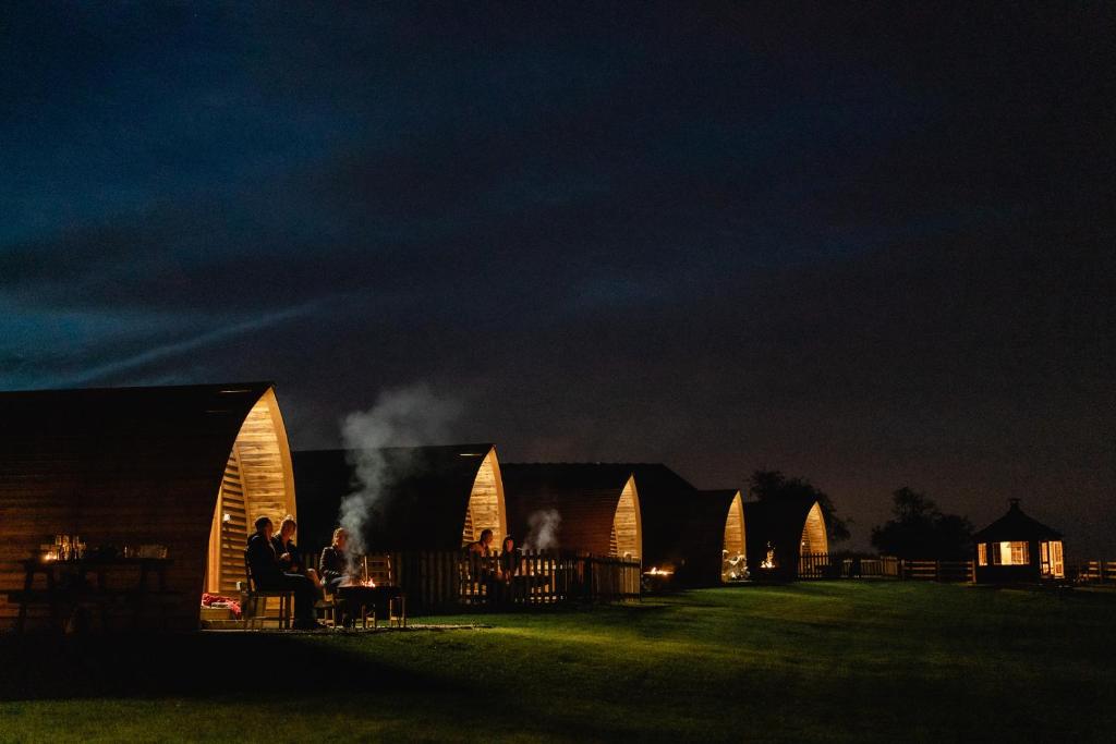 a group of people sitting at a table in front of wooden buildings at night at Saxon Meadow by Wigwam Holidays in Tamworth