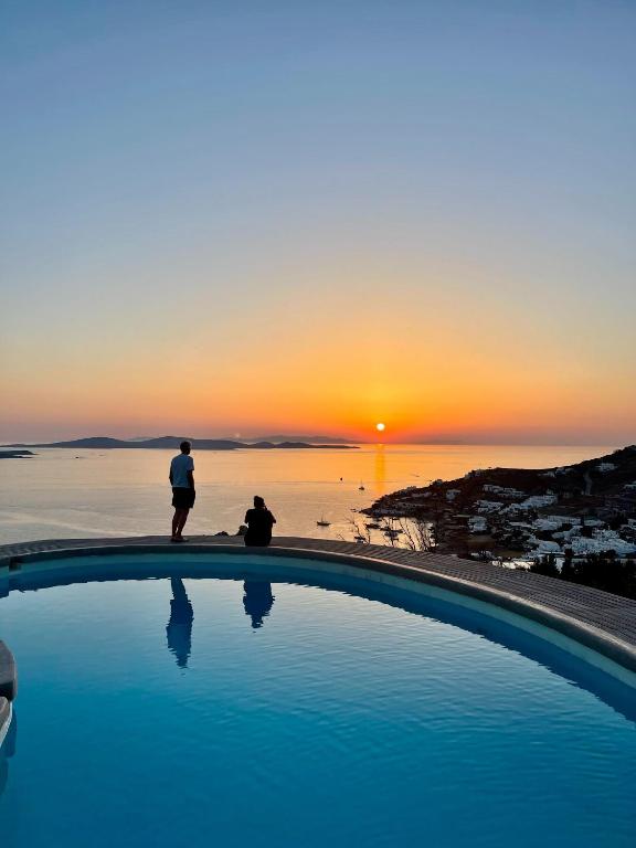 two people standing next to a swimming pool at sunset at Villa Tavani Agios Ioannis Mykonos in Agios Ioannis Mykonos