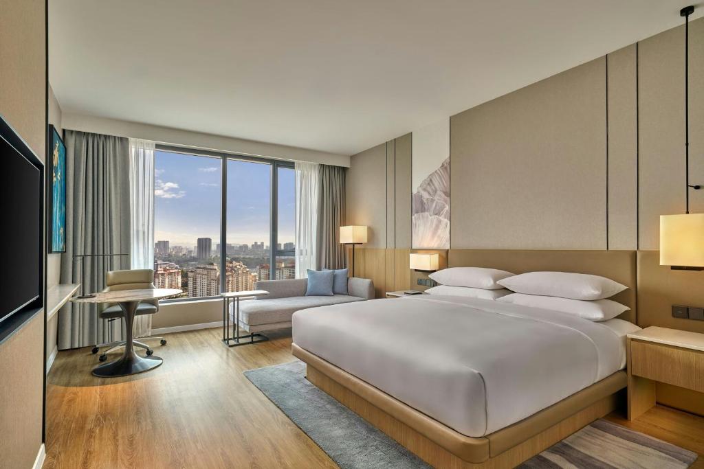 A spacious bedroom with a large bed, sofa seating, desk and panoramic city-facing window.