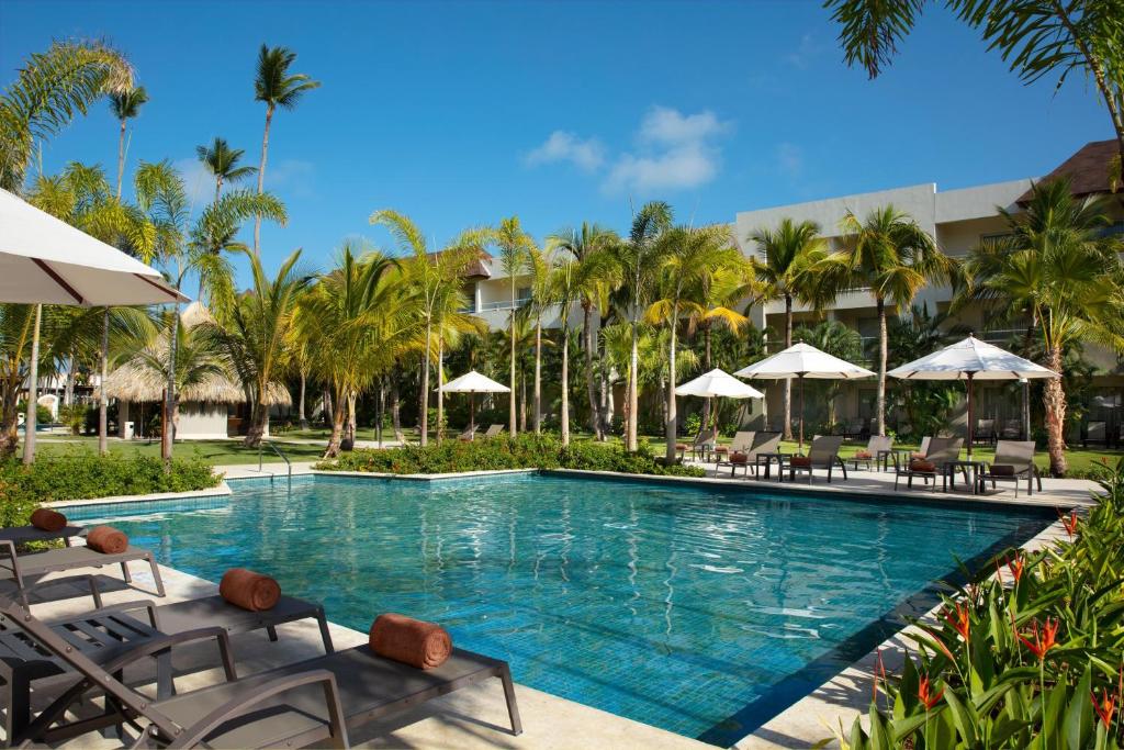 Resort pool with lounge chairs, white umbrellas, palm trees, and modern building backdrop.