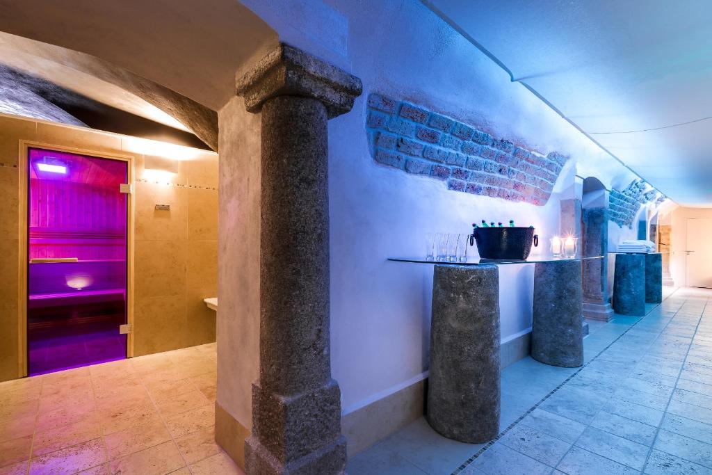Dim spa corridor with stone columns, glass-tiled floor, purple-lit sauna doorway, chilled bottles on glass table.