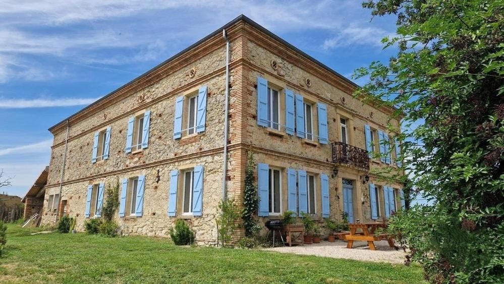 un grand bâtiment en briques avec des fenêtres bleues sur un champ dans l'établissement Aux roses de Cèdres chambre Savane, à Salvagnac