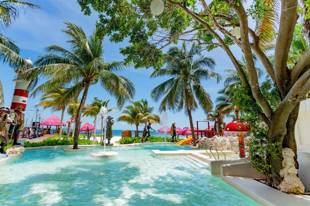 Water park area with shallow pools, water slide, palm trees, and pink umbrellas by beach.