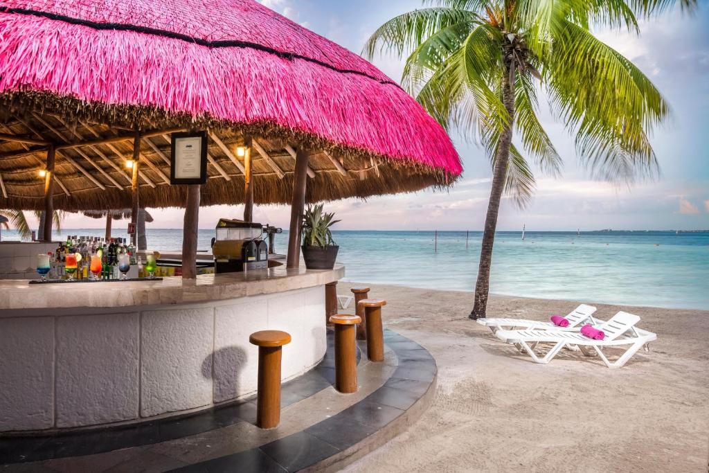 Beachfront bar with pink umbrella, wooden stools, and ocean view behind.