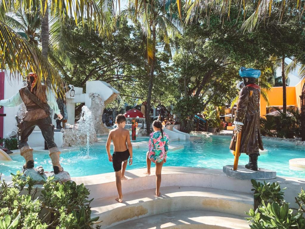 Children running toward turquoise pool with pirate statues and tropical landscaping.