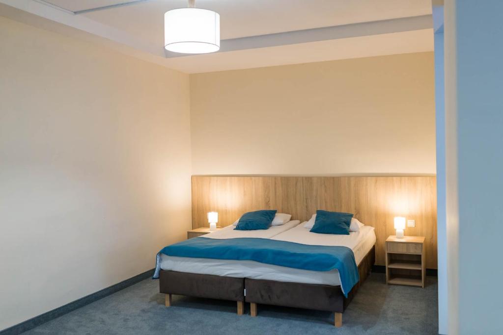 Simple hotel bedroom with double bed, teal pillows and blanket, bedside lamps, neutral walls.