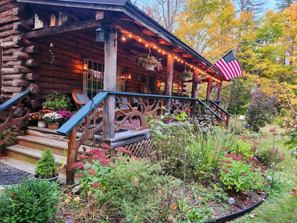a log cabin with a porch with flowers and an american flag at Deer Valley Retreat, Lovely Log Cabin with Fireplace & Garden Views- Perfect for a Relaxing Getaway! in Bradford