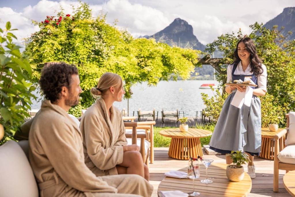 a woman talking to two people sitting on a deck next to a lake at Hotel Seevilla Wolfgangsee in St. Wolfgang