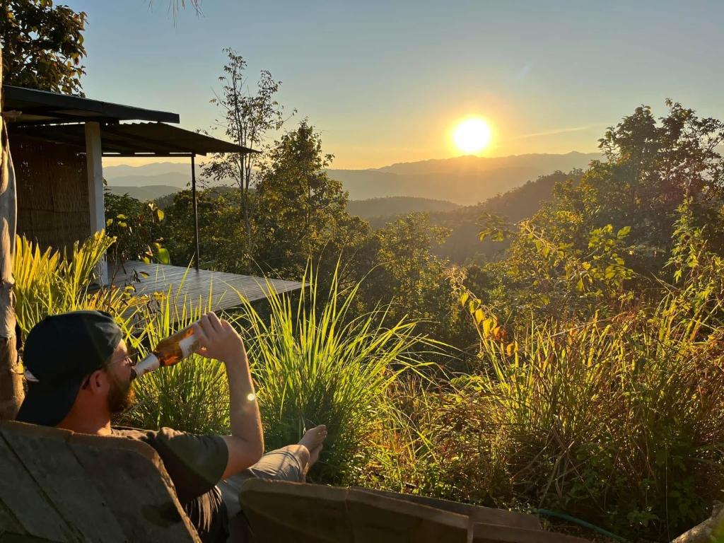 a man is sitting on a bench drinking a drink at Monsonkeang Camping view ม่อนสันเกี๋ยง in Mae Chaem