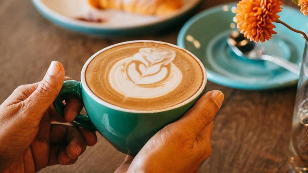 A latte with foam art is served in a turquoise cup beside a pastry and flower.