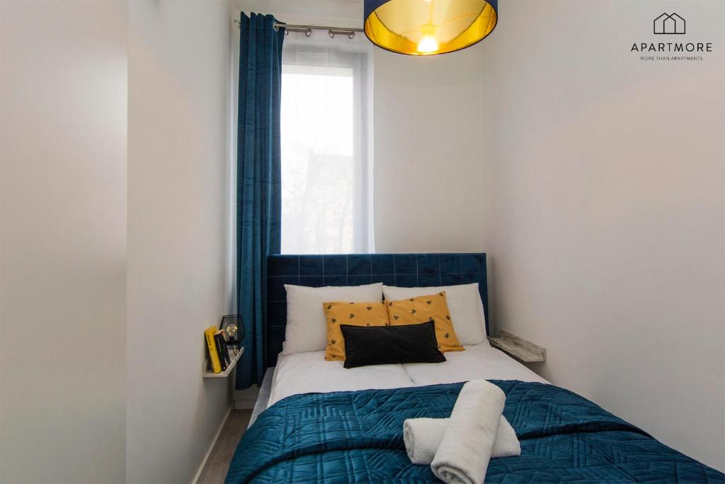 Compact bedroom with teal quilted bedspread, mustard pillows, tall window with curtain, rolled towels.