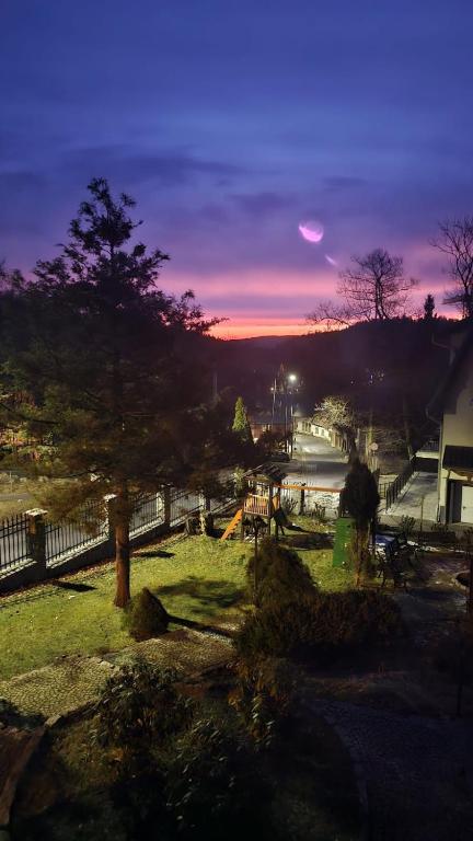 Twilight view over garden and playground, street lamps lit, purple-pink sky with crescent moon.