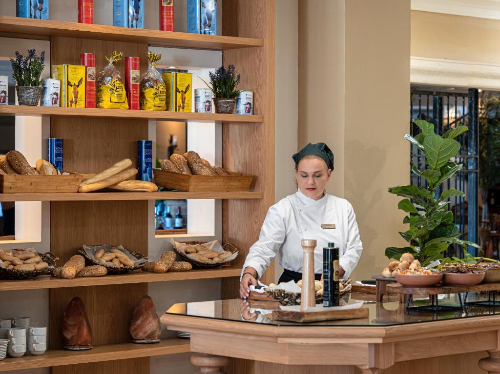 a woman in a kitchen with a table with bread at Grecotel Marine Palace & Aqua Park in Panormos Rethymno