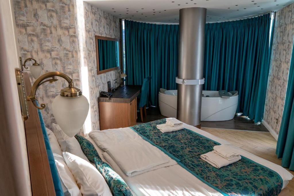 King bed with patterned runner and folded towels, brass wall sconce, desk, floor-to-ceiling teal curtains and freestanding tub at City Palace Hotel in Ohrid