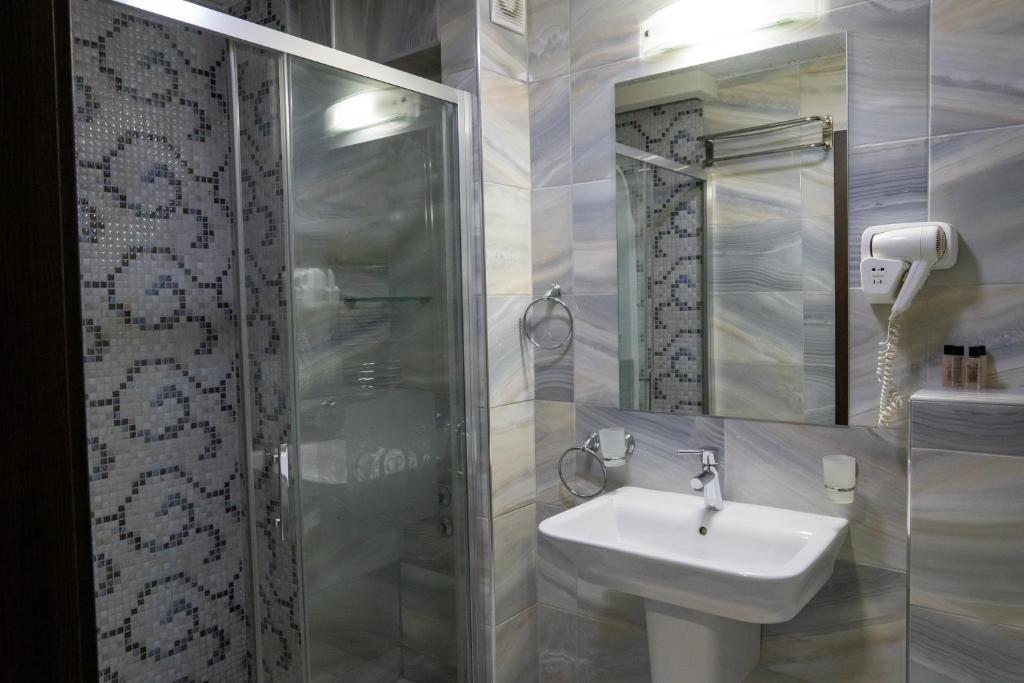 Glass shower with patterned mosaic accent, wall-mounted hairdryer, sink with soap dispenser and towel rack on blue-gray tiles at City Palace Hotel in Ohrid