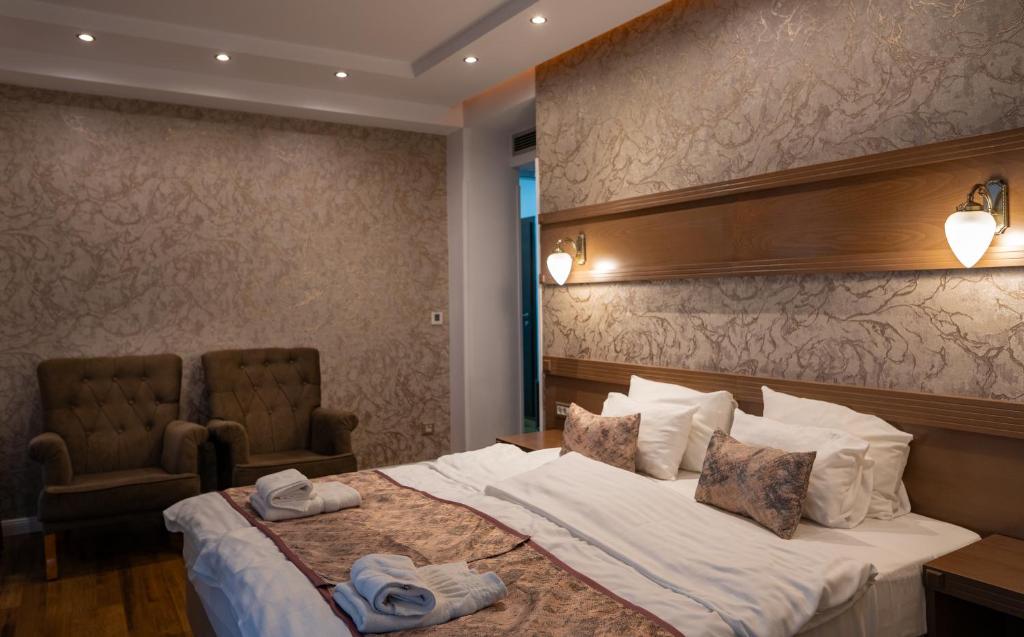 Double bed with patterned runner and pillows, upholstered armchairs, wooden headboard with wall sconces and rolled towels at City Palace Hotel in Ohrid