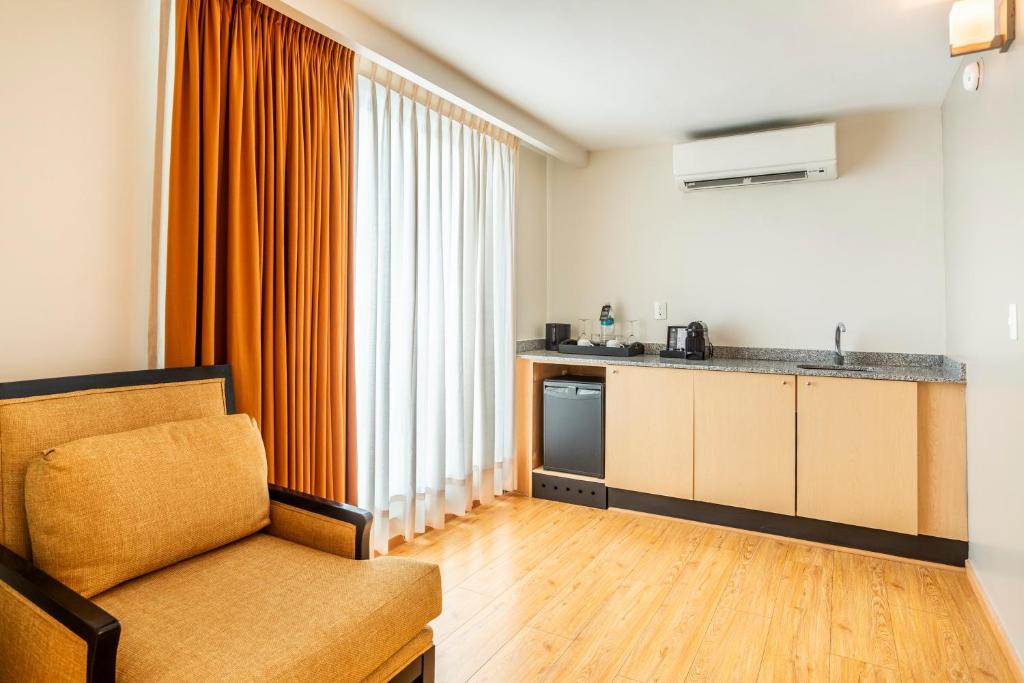A lounge chair faces a compact kitchenette beside tall curtained windows.