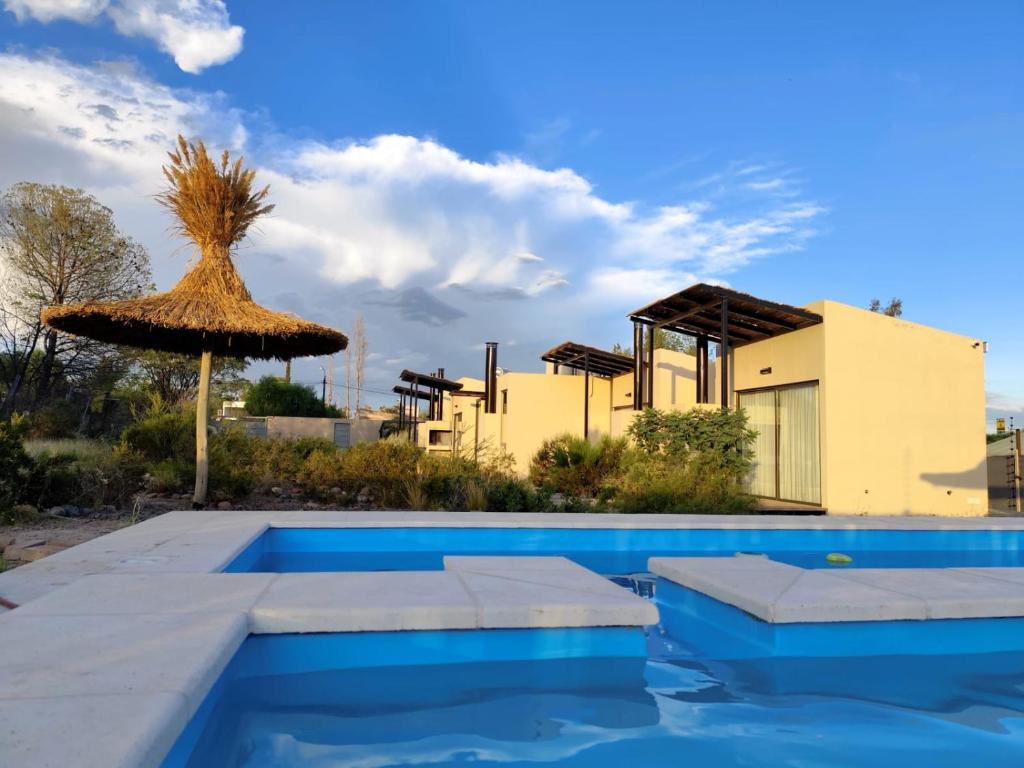 a swimming pool with a straw umbrella and a house at Posada sustentable Los Chañares con vista a la montaña in Ciudad Lujan de Cuyo