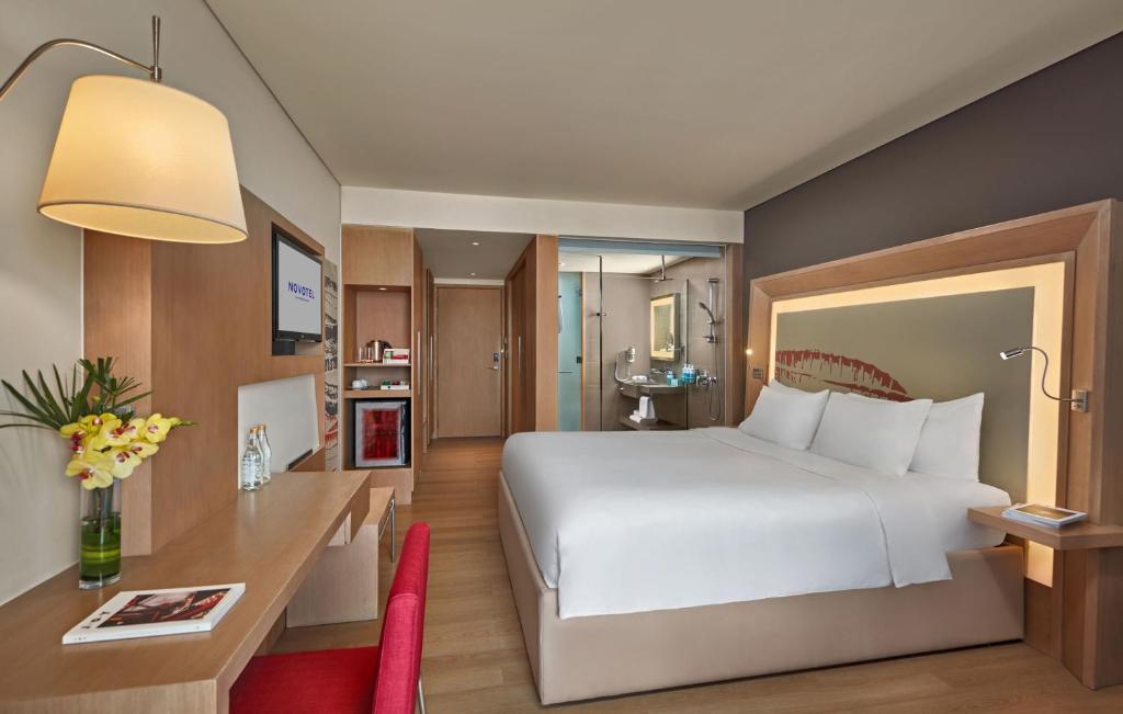 A bright guest room with a large white bed, built-in desk, minibar and glass-enclosed bathroom.