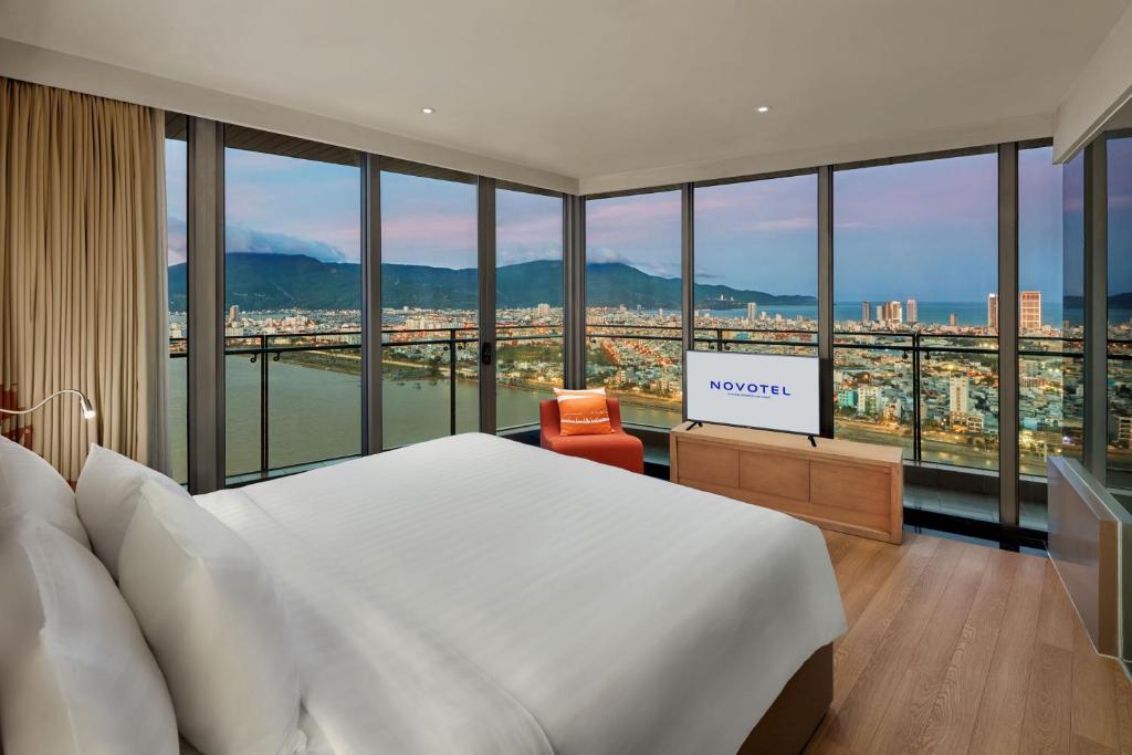 A bright hotel bedroom with a king-size bed and floor-to-ceiling windows overlooking the river, city, mountains and coast.