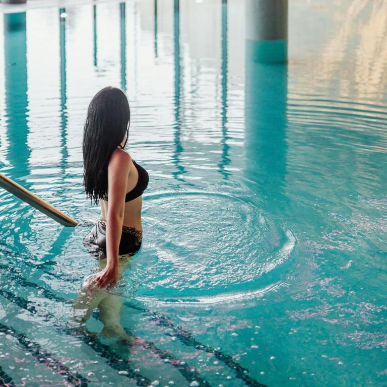 Woman wading into indoor pool using handrail, gentle ripples spreading around her.