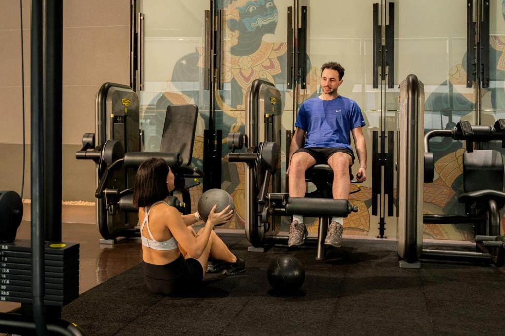 A compact, equipment-filled gym with strength machines, free weights, and two guests exercising.