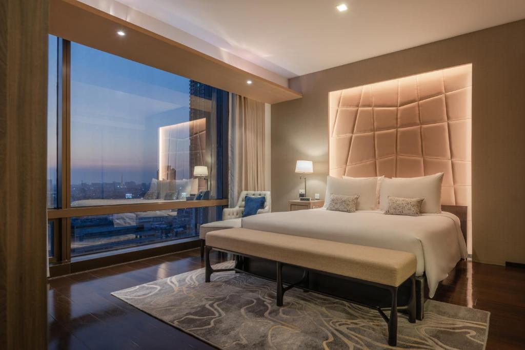 An upscale bedroom with a padded illuminated headboard, city-facing window, and upholstered bench.