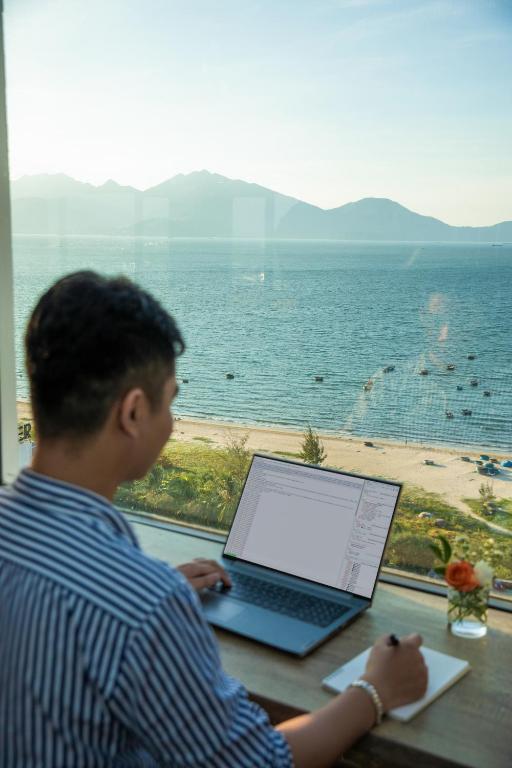 A window-side work setting with a laptop, notebook, and direct beach-and-sea view.