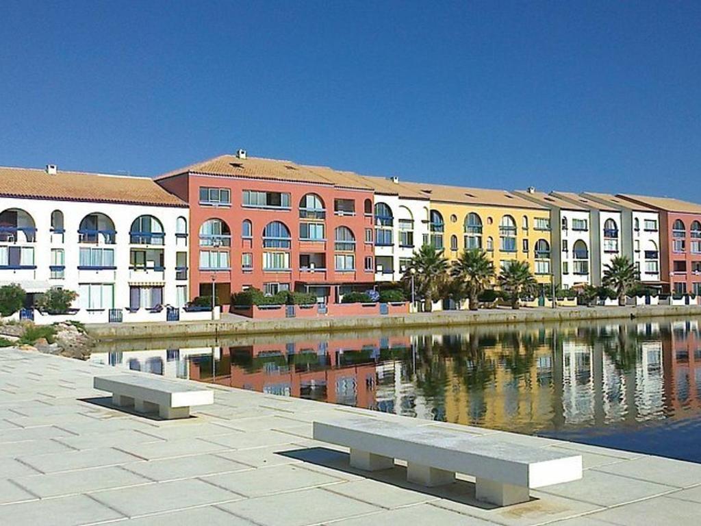 un bâtiment avec un banc devant un plan d'eau dans l'établissement Appartement 2P avec Clim, Terrasse et Parking, Le Barcarès - FR-1-782-76, au Barcarès