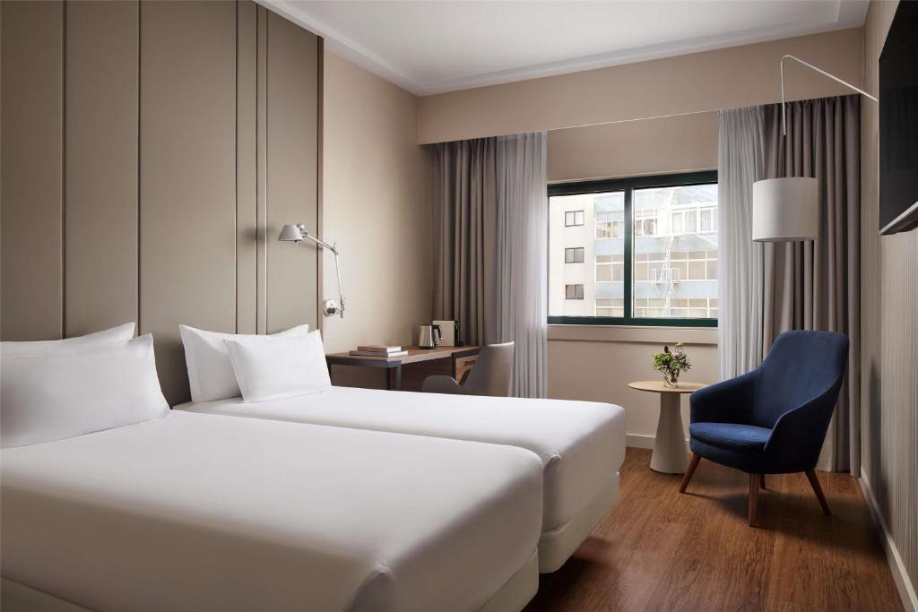 A bright twin room with two single beds, a blue armchair and a city-facing window.