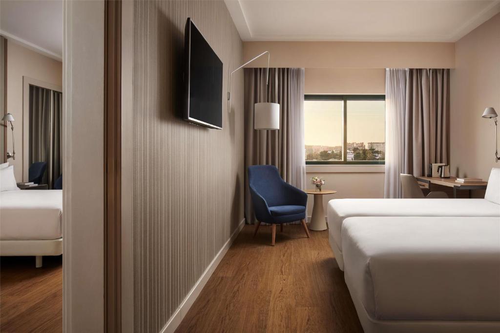 A modern hotel room with a bed, desk, armchair and a broad urban view through the window.