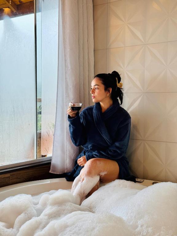 a woman sitting on a bed holding a cup of coffee at Chalés Vila do Moinho in Monte Verde