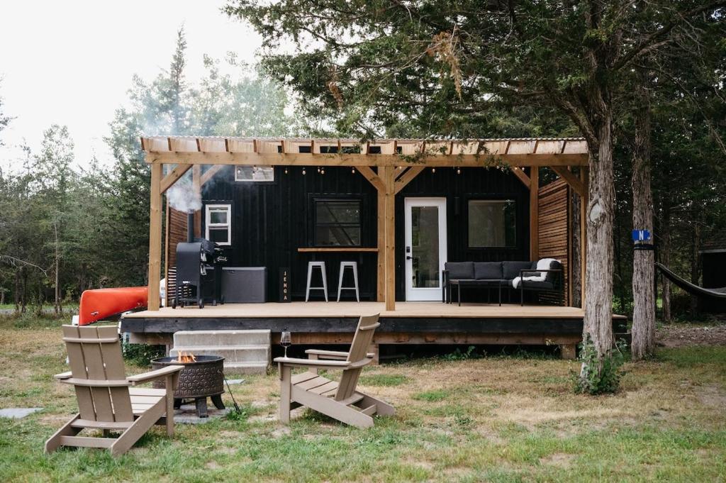 a black tiny house with a porch and two chairs at The Loft - Stargazing Tiny Home with Firepit, Wood Fired Pizza Oven & Movie Projector in Deseronto