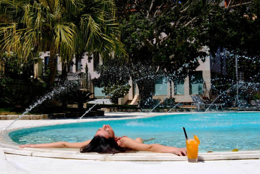 una donna sdraiata in piscina con un drink di Hotel Parco delle Fontane a Siracusa