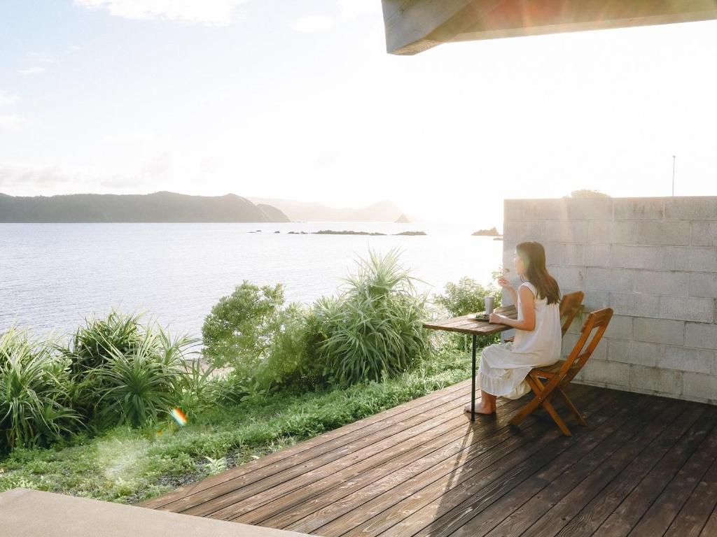 a woman sitting at a table on a deck overlooking the water at Denpaku The Beachfront MIJORA in Amami