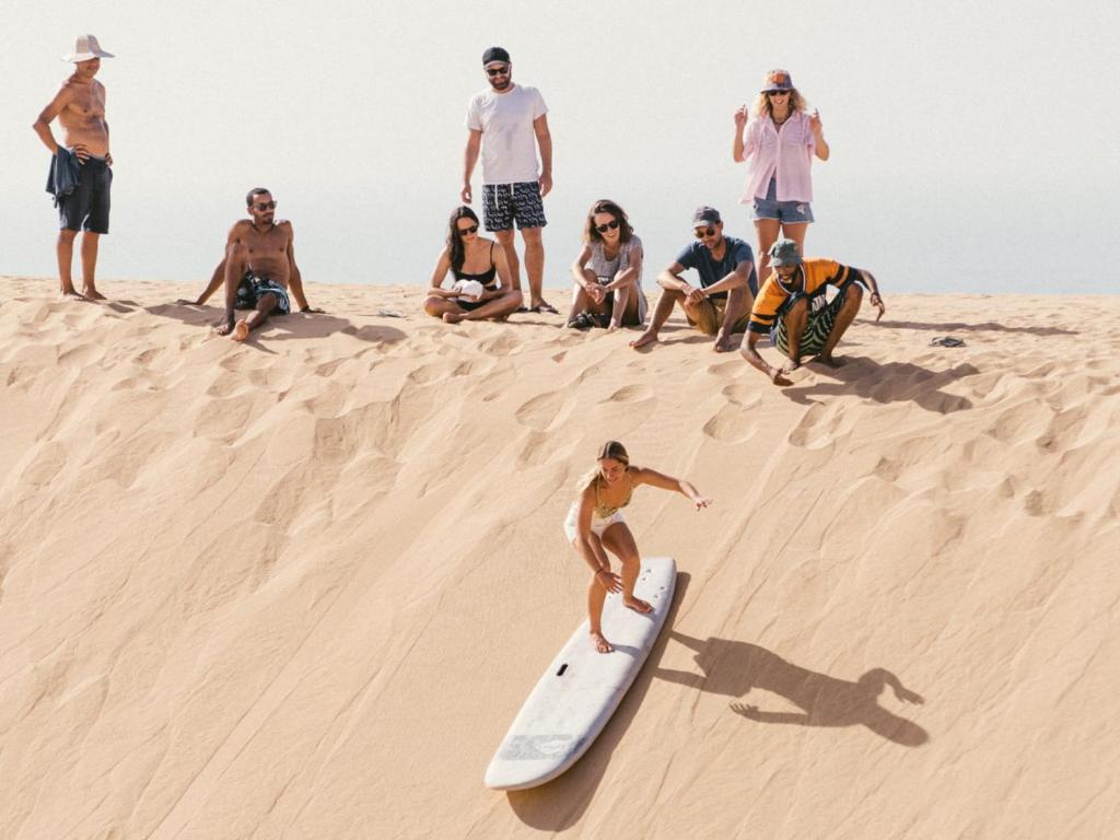 a group of people sitting on the sand with a girl on a surfboard at Tamraght Surf House in Tamraght Ouzdar